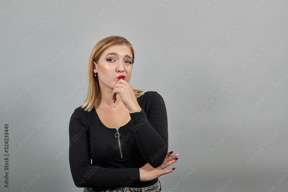 Young blonde girl in black jacket on grey background dreamy woman doubts, holds hand to face Young blonde girl in black jacket on grey background dreamy woman doubts, holds hand to face