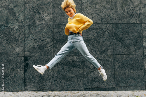 Positive young woman wearing yellow sweater, blue hight waist jeans, jumping high, feeling happy on the city street. Student blonde female as joyful expression