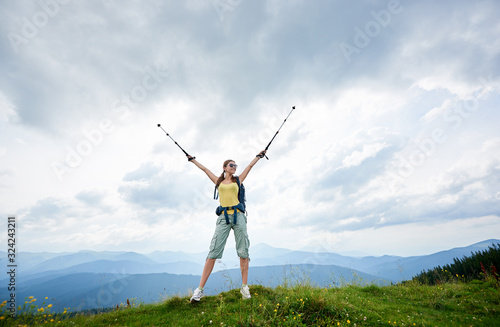 Happy woman hiker hiking mountain trail, standing on a top of grassy hill with raised hands, holding trekking sticks, wearing backpack and sunglasses, enjoying summer day. Outdoor activity concept