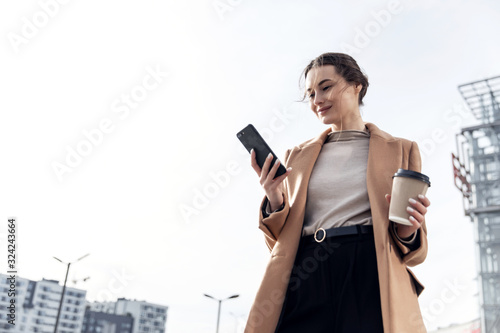 Closeup young Business Woman holding smartphone, Coffee to go and smiling. Attractive stylish Woman using her Phone near modern Building. Having Break. Smartphones. Games. People. Emotions. Business.