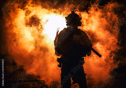 Fotografija Silhouette of modern infantry soldier, elite army fighter in tactical ammunition and helmet, standing with assault service rifle in hands on background of fiery explode
