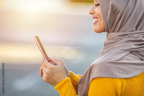 Female in her 20s wearing hijab texting on cellphone on urban street. Young woman wearing hijab head scarf in city texting cell phone. Portrait of happy muslim woman using mobile phone