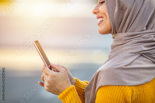 Woman wearing hijab browsing internet on mobile phone outdoors. Portrait of a modern arabian girl using mobile phone on the street. Close up of beautiful woman wearing hijab smiling while texting 
