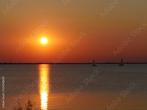 Breathtaking sunset at the lake with rays reflected in the water, with sailboats in the distance.