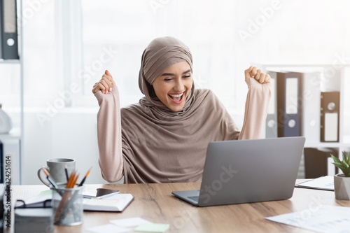 Happy muslim female office employee looking at laptop screen, celebrating success