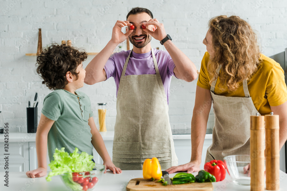 happy and bearded homosexual man covering eyes with cherry tomatoes near mixed race son and partner happy and bearded homosexual man covering eyes with cherry tomatoes near mixed race son and partner