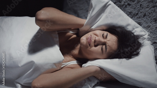 Photography Portrait of young woman lying in bed and closing ears with pillow suffering from