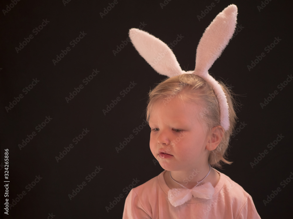 Funny blond child in pink pajamas and easter bunny costume eats carrot. Portrait of boy on black background