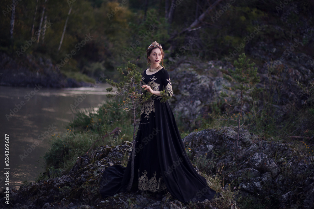 A girl in a black dress with a crown on her head stands on the banks of the river