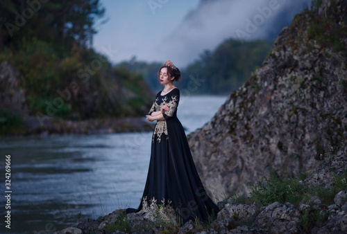 A girl in a black dress with a crown on her head stands on the banks of the river