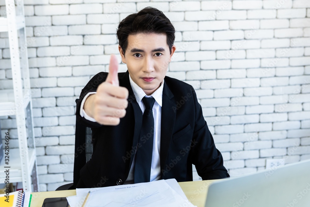 Happy mood a cheerful successful asian young businessman worked with laptop computer positive adult business showing thumbs up and ideas on wooden table background in office Happy mood a cheerful successful asian young businessman worked with laptop computer positive adult business showing thumbs up and ideas on wooden table background in office