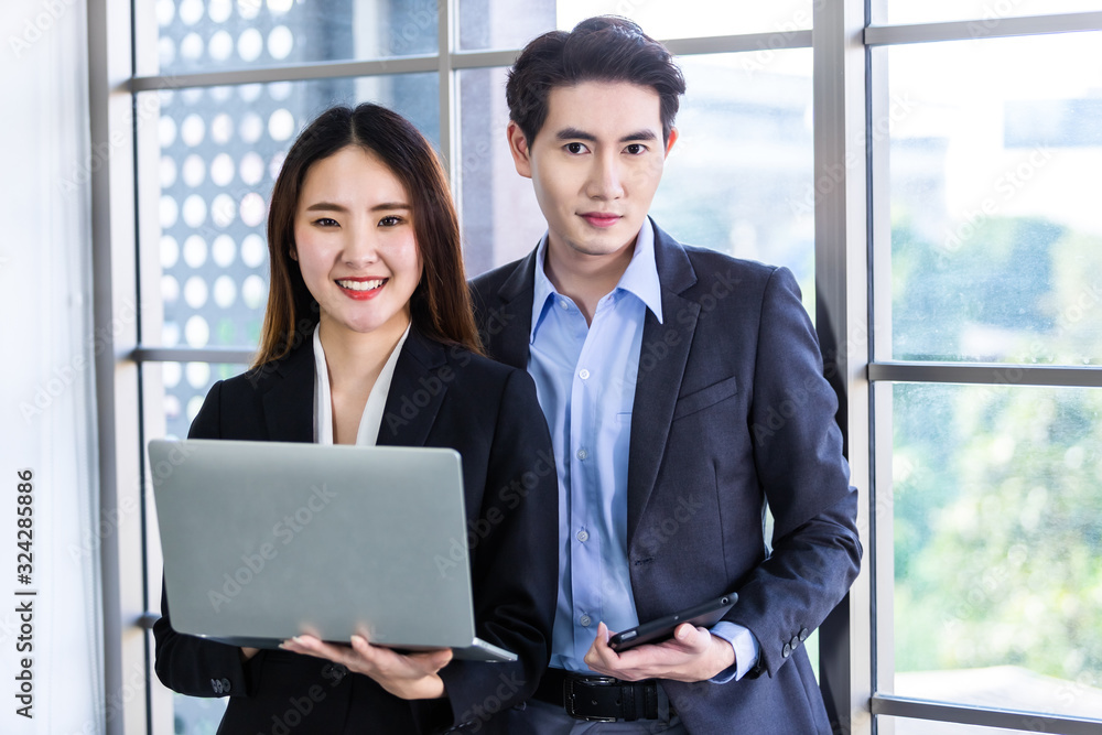 Young Asian businesswoman and businessman partners while working together with laptop computer and business man holding a tablet in office background,Working couple Young Asian businesswoman and businessman partners while working together with laptop computer and business man holding a tablet in office background,Working couple