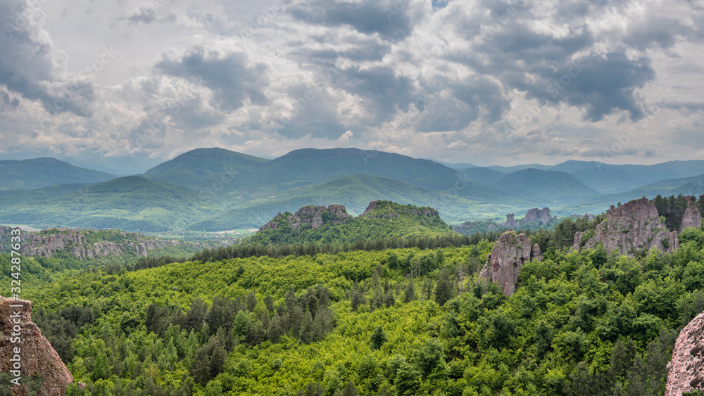 Panoramic view of a green mountain valley with dense forests and scattered large rock formations, Belogradchik Rocks, Bulgaria. Mountains and dramatic cloudy sky in the background.