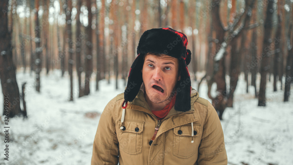 Man grimaces in forest in winter season. Portrait of crazy male wearing earflap hat and grimacing face close up in winter forest. Man grimaces in forest in winter season. Portrait of crazy male wearing earflap hat and grimacing face close up in winter forest.