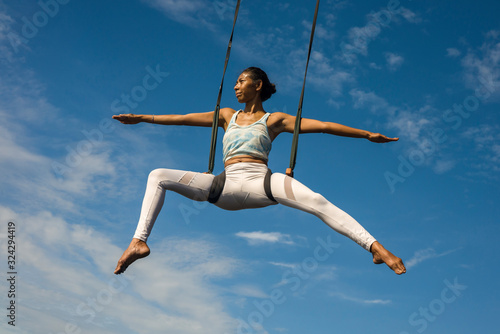 outdoors portrait of young happy and athletic Asian Indonesian woman doing aero yoga workout training body balance hanging from swing rope isolated on blue sky