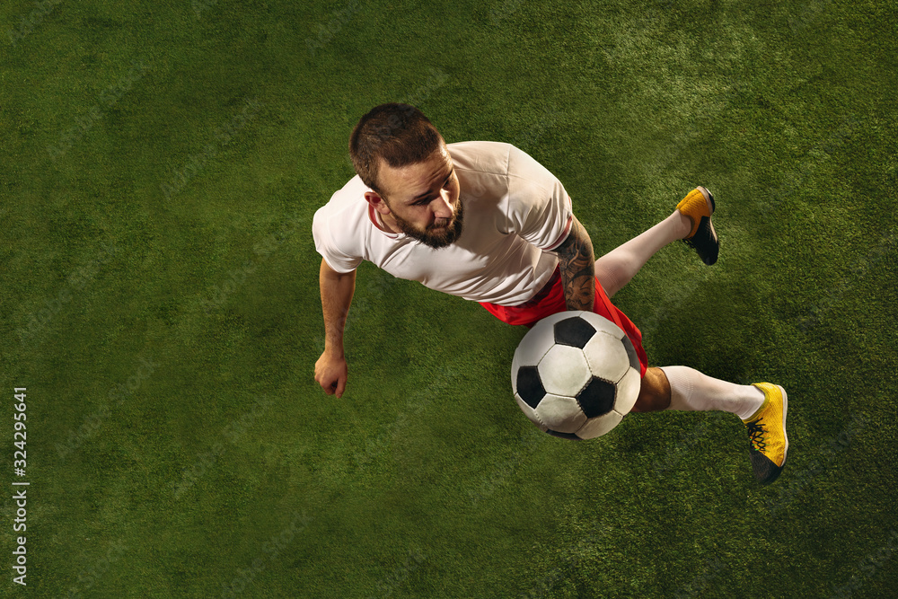 Top view of caucasian football or soccer player on green background of grass. Young male sportive model training, practicing. Kicking ball, attacking, catching. Concept of sport, competition, winning.