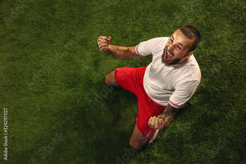 Top view of caucasian football or soccer player on green background of grass. Young male sportive model training, practicing. Kicking ball, attacking, catching. Concept of sport, competition, winning. photo