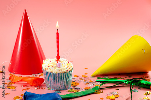 Happy Birthday and surprise party concept with frosted cupcake and lit candle, party hats, confetti and balloons on pink background with copy space
