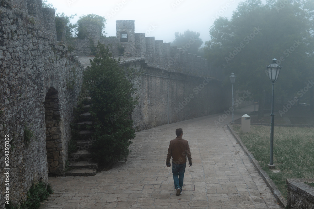 Man walking alone at distance. Fortification on top of the mountain, old castle. Journey. Mystical atmosphere, fog, white haze, mist. Summer day.