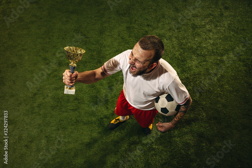 Top view of caucasian football or soccer player on green background of grass. Young male sportive model celebrating win with champions cup, emotional screaming. Concept of sport, competition, winning.