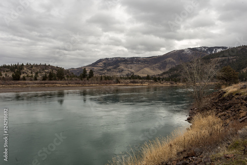 Calm green river in front of tree covered rolling hills on an overcast day
