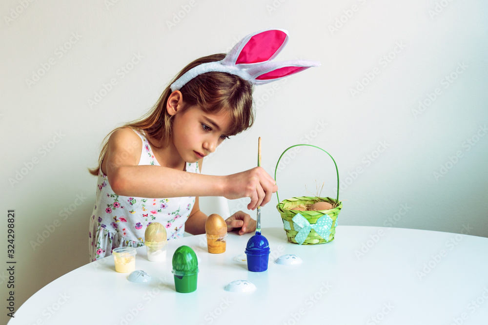 Beautiful eight-year-old girl with bunny ears paints eggs at a white table. Easter concept. Beautiful eight-year-old girl with bunny ears paints eggs at a white table. Easter concept.