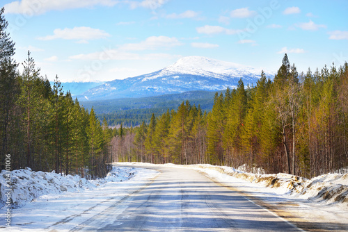 A view of the empty asphalt road through the coniferous forest. Snow-covered mountains in the background. Winter landscape. Kola Peninsula, Murmansk region, Polar Circle, Karelia, Russia photo