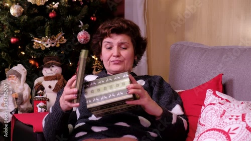 Christmas Gift - Sad woman lying on a sofa near the christmas tree, opening gift disappointed and unhappy. concept of holidays and new year.