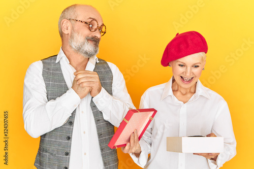 Billede på lærred Studio image of emotional elegant male pensioner with gray beard and bald head looking with impatience, watching attractive woman opening box with present from him