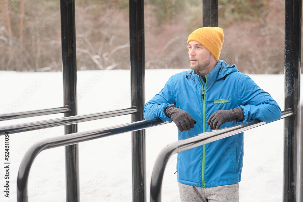 Serious young man with stubble standing between parallel bars on sports ground in winter and looking into distance Serious young man with stubble standing between parallel bars on sports ground in winter and looking into distance