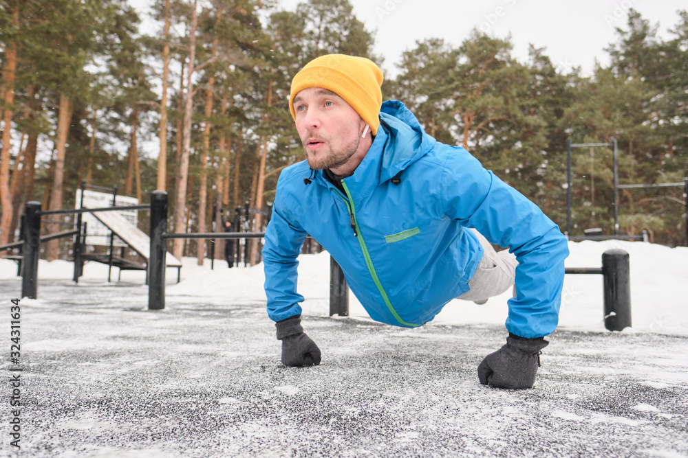 Strong young man in ear buds pushing himself away from ground while training at workout area in winter Strong young man in ear buds pushing himself away from ground while training at workout area in winter