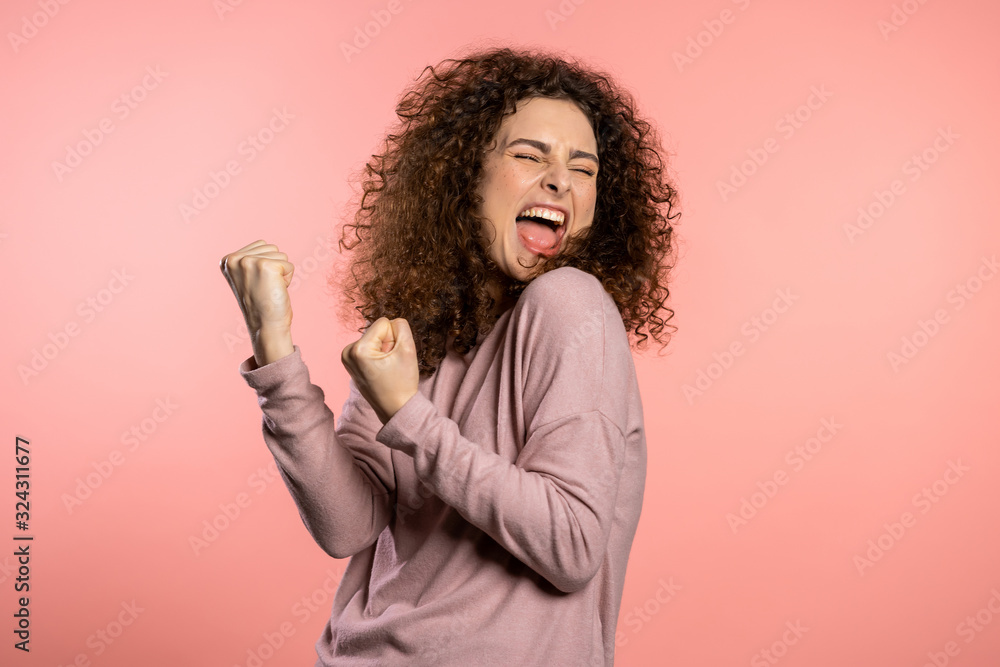 Girl with curly hair and extraordinary makeup shows yes gesture of victory, she achieved result, goals. Surprised excited happy woman on pink background Girl with curly hair and extraordinary makeup shows yes gesture of victory, she achieved result, goals. Surprised excited happy woman on pink background