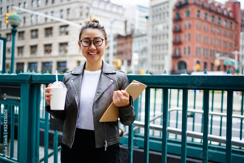 Half length portrait of cheerful female student with sketchbook and takeaway milk shake in hands standing at urbanity area and smiling at camera, happy Asian woman in tredny spectacles posing outdoors photo