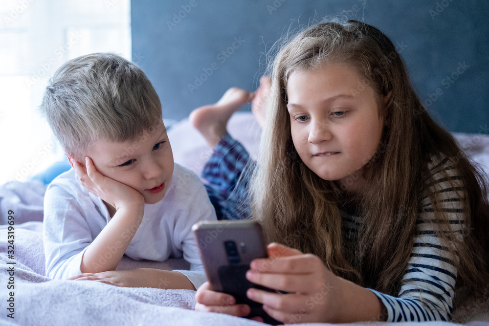 Two kids caucasian boy and girl playing games or looking information video text at Internet on mobile phones at home lying in bed. Digital technology, Modern childhood . Two kids caucasian boy and girl playing games or looking information video text at Internet on mobile phones at home lying in bed. Digital technology, Modern childhood .