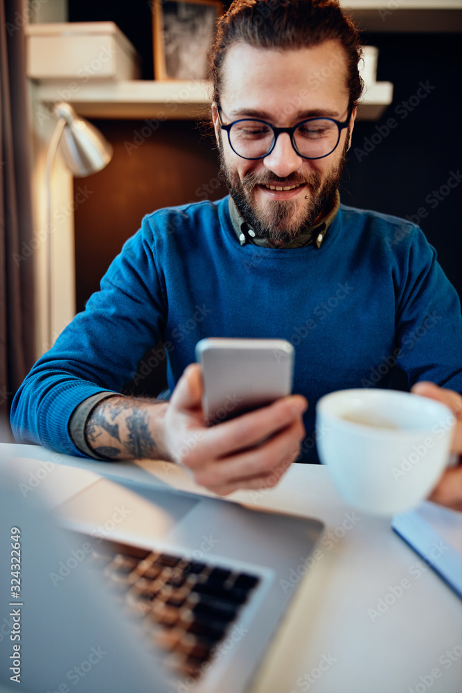 Young handsome bearded hipster sitting at his home office, holding cup of coffee and using smart phone. Young handsome bearded hipster sitting at his home office, holding cup of coffee and using smart phone.
