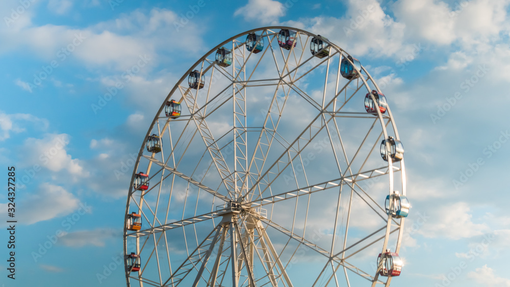 White clouds and rotating ferris wheel at festival or carnival. Summer cloudy day, warm illumination, sunset blue sky. Sightseeing, cityscape, entertainment, holiday, leisure time concept