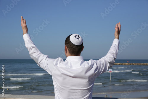 young Jew man in a kip pah hat is standing with his back with raised hands praying against the blue sea and blue sky.