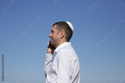 Obraz na plátně portrait of a young jew in kippah talking on a cell phone on a blue sky background