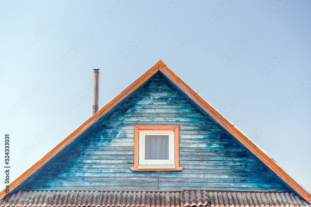 Fragment of facade of wooden house with cracked weathered blue paint, with iron pipe on roof and plastic window with white frame against sunny sky. Rustic style
