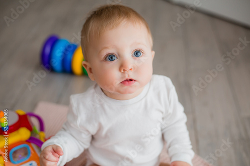 baby in white clothes is sitting on the floor in the children's room and playing with toys