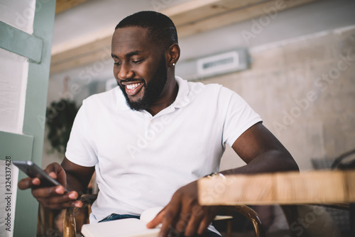 Joyful young black male student messaging on smartphone in cafe