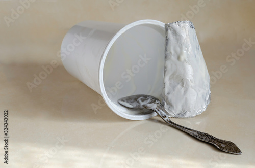 Empty plastic cup of yogurt. Spoon with the remains of the dairy product inside the fallen glass. Kitchen table after breakfast. Eye level shooting. Selective focus.