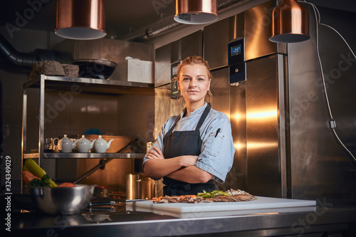 Confident and serious female chef standing with hands crossed in a dark kitchen next to cutting board with vegetables on it, wearing apron and denim shirt, looking in the camera, cooking show look photo