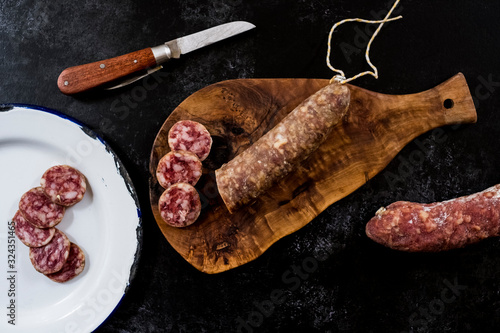 High angle close up of knife, sliced salami on wooden cutting board and white enamel plate on black background. photo