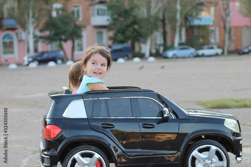 Two amazing girls ride in one big toy car on city street asphalt. Outdoor driving in a summer attraction for children Two amazing girls ride in one big toy car on city street asphalt. Outdoor driving in a summer attraction for children