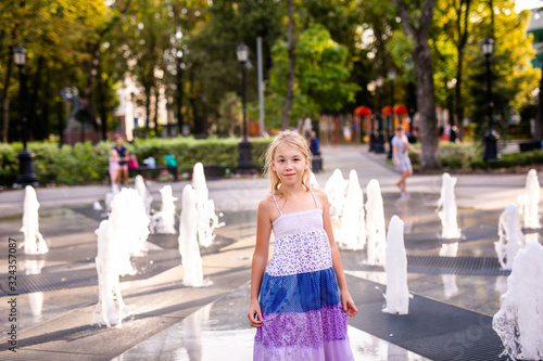 Blonde happy child girl in long purple dress running between water flow in city summer park. Child activity concept