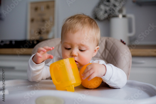 baby is sitting in a high chair, holding an orange. a baby in a high chair drinks water from an orange drinking bowl