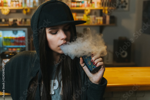 Young pretty woman in cap smoke an electronic cigarette at the vape shop. Hip-hop style. Closeup. photo