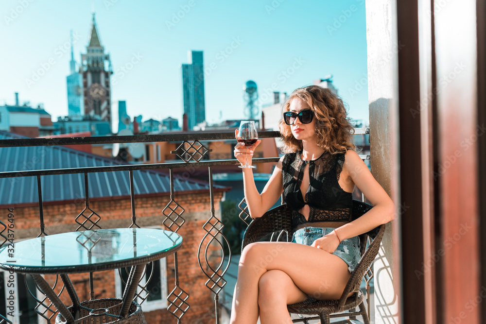 Pretty girl in a black bodysuit with a glass of wine sits on a balcony against the background of  Europe city view landscape. 
