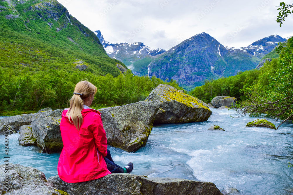 Naklejka premium The young woman near river which is located near path to the Briksdalsbreen (Briksdal) glacier. The melting of this glacier forms waterfall and river with clear water. Jostedalsbreen National Park. No